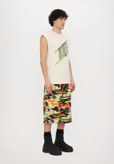 Beige ermeløs t-skjorte med "BLUE" grafikk, kombinert med knelange camo shorts i grønn, oransje og svart; svarte kraftige støvler.