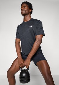 Zwart sportshirt met een subtiel patroon, logo op de borst. Houdt een zwarte kettlebell vast. Sportshorts in het zwart. Neutrale achtergrond.