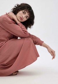 Abito lungo e rosa con una silhouette fluida, realizzato in tessuto morbido. La modella si accovaccia, mostrando la lunghezza completa dell'abito e il suo design semplice ed elegante.