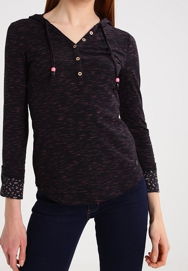 Frau mit einem schwarzen Hoodie mit langen Ärmeln, der pinke Sprenkel, Holzknöpfe und Blumenmuster an den Bündchen hat, kombiniert mit dunkelblauen Jeans.