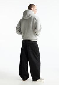 Felpa grigia con cappuccio e vestibilità rilassata, abbinata a jeans neri a gamba larga e sneaker grigie su uno sfondo chiaro.