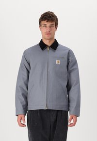 Veste en toile grise avec col en velours côtelé noir, fermeture éclair à l'avant et poches latérales ; présente un écusson de marque sur la poitrine.