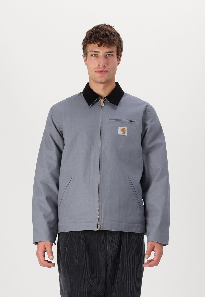 Veste en toile grise avec col en velours côtelé noir, fermeture éclair à l'avant et poches latérales ; présente un écusson de marque sur la poitrine.
