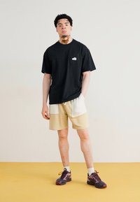 Svart kortermet t-skjorte med lomme foran, kombinert med beige shorts med hvite detaljer og to sidelommer. Brukt med flerfargede joggesko.