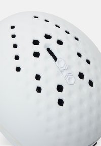 Biały kask z heksagonalnymi otworami wentylacyjnymi, gładką teksturą i regulowanym czarnym paskiem. Centralna wypustka z detalem mechanizmu blokującego.