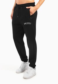 Schwarze Jogginghosen mit elastischem Bund, Seitentaschen und weißer "SMILODON"-Stickerei auf dem Oberschenkel, getragen mit weißen Sneakern.