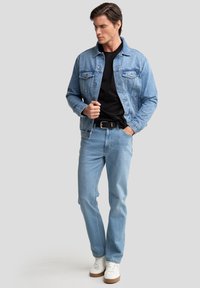 Hellblaues Denim-Jackett mit zwei Brusttaschen, getragen über einem schwarzen Shirt, kombiniert mit hellen Jeans und weißen Sneakers.