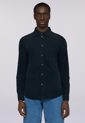 Hombre vestido con una camisa de botones azul marino oscuro con un bolsillo en el pecho y jeans de mezclilla claros, de pie contra un fondo liso claro.