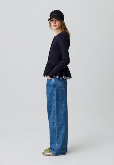 Giovane donna con maglione nero a peplo, jeans blu a gamba larga, scarpe argento con lacci gialli e cappellino nero, in piedi di lato con le mani nelle tasche.