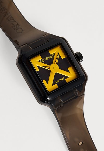 Montre carrée Off-White noire et jaune avec de grandes flèches jaunes et un bracelet translucide marron foncé sur fond blanc.