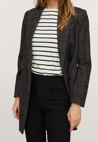 Blazer à carreaux sombres en tissu texturé, présentant une silhouette structurée, des revers crantés, des poches latérales et une fermeture à bouton. Associé à un t-shirt rayé blanc et marine et un pantalon noir taille haute.