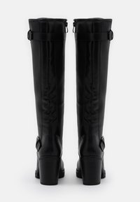 Botas altas de cuero negro con tacones de bloque y correas ajustables en la parte superior y cerca del tobillo, vistas desde atrás sobre un fondo blanco.