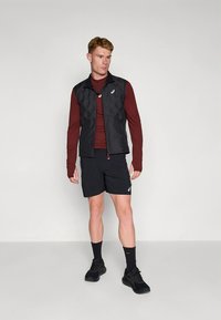 Schwarze Steppweste über einem dunkelroten Langarmshirt, kombiniert mit schwarzen Shorts und schwarzen Sportschuhen. Glatte Textur, minimalistisches Design.