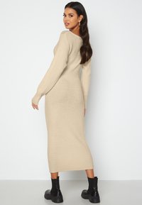 Abito in maglia a costine beige con maniche lunghe e silhouette aderente, che termina sotto il ginocchio. Abbinato a stivali neri alla caviglia.