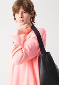 Schwarze Leder-Tote-Bag mit strukturierter Form, die eine glatte Textur und einen breiten Trageriemen aufweist. Modell trägt ein leuchtend pinkes Langarmoberteil.