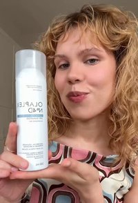 Jonge vrouw met krullend blond haar houdt een witte fles Olaplex No. 4D Clean Volume Detox Droogshampoo vast en presenteert deze binnen.