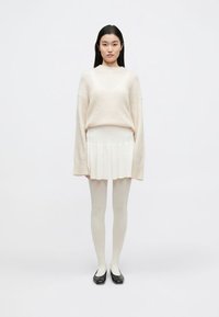 Pulover supradimensionat bej deschis, fustă mini plisată albă și colanți albi transparenți. Pantofi negri comozi completează ținuta. Design simplu, minimal.