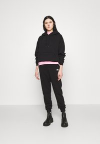 Sort hætte sweatshirt med en frontlomme, lagdelt over en pink skjorte. Sorte sweatpants med et accent logo, parret med robuste sorte støvler.