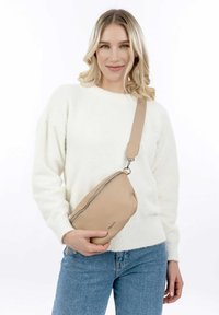 Béžová kožená kabelka přes rameno s texturovaným povrchem, zapínáním na zip a nastavitelným popruhem, držena proti bílé svetru a modrým džínům.