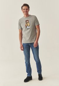 Szürke póló egy sárga kabátos medve grafikával; kék farmerrel és fekete csizmával párosítva. Egyszerű dizájn, puha textúra és kényelmes fazon.