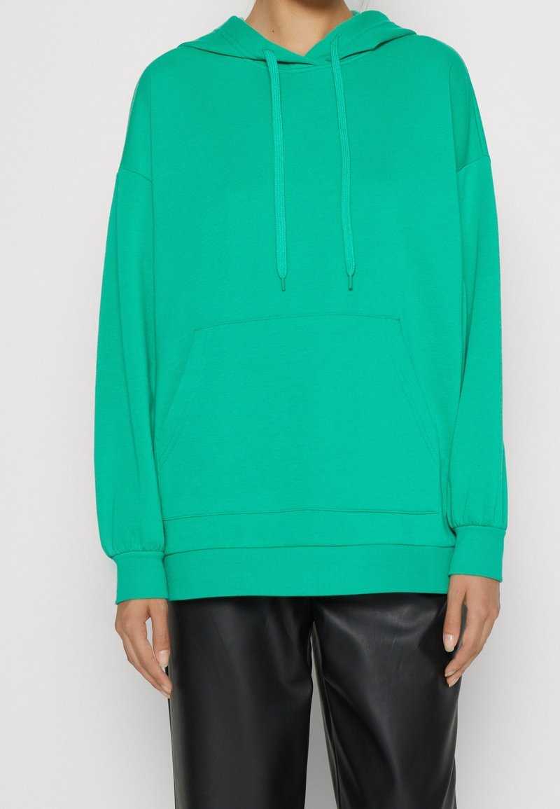 Personne portant un sweat à capuche bleu sarcelle avec poche avant et cordons, associé à un pantalon en cuir noir sur un fond uni.
