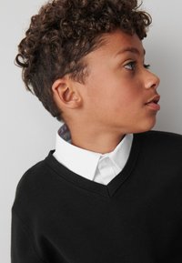 Črn pulover z v-izrezom, oblečen preko bele srajce z ovratnikom, teksturirane tkanine, z gladkim ovratnikom in subtilnim vzorcem, ki je viden na izrezu.