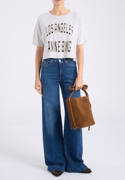 Άτομο που φορά cropped γκρι μπλουζάκι "Los Angeles Anne Bing", φαρδιά μπλε τζιν, κρατώντας καφέ σουέτ τσάντα με χερούλι φιόγκο.