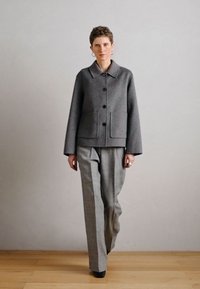 Γκρι μάλλινο παλτό με γιακά, μπροστινές τσέπες και κουμπώματα, σε συνδυασμό με ανοιχτό γκρι φαρδιά παντελόνια. Το μοντέλο στέκεται σε ξύλινο πάτωμα.