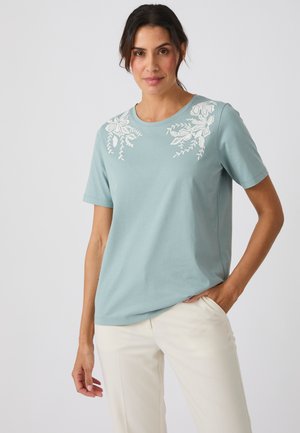 Femme portant une chemise à manches courtes bleu clair avec une broderie florale blanche sur les épaules et un pantalon couleur crème, debout devant un fond uni.