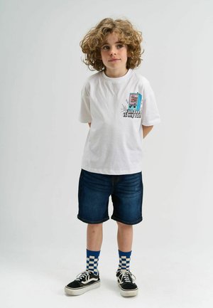 T-shirt blanche à manches courtes avec un motif graphique d'un distributeur automatique sur le devant. Portée avec un short en denim foncé et des baskets noires avec des chaussettes à carreaux bleus.