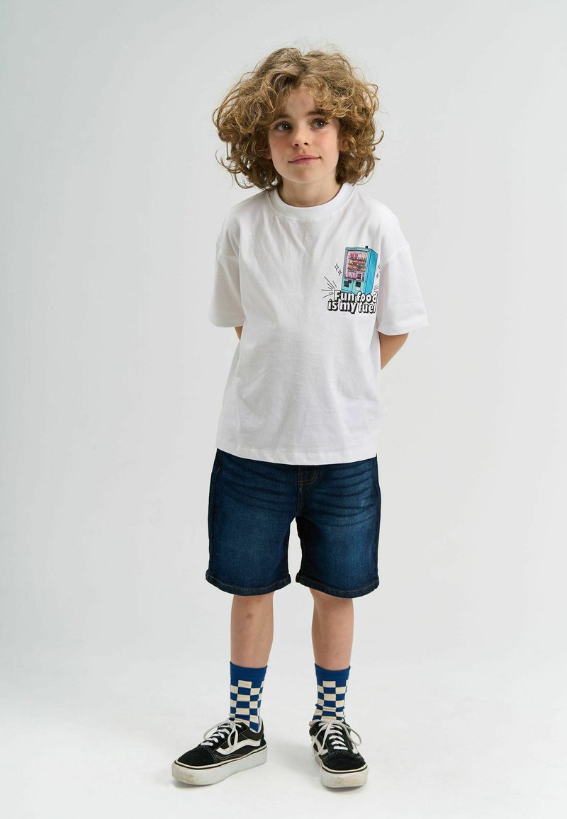 Bijela majica s kratkim rukavima sa grafičkim uzorkom automata za prodaju sprijeda. U kombinaciji s tamnim trapericama i crnim tenisicama uz plave karirane čarape.