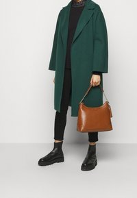 Grön ullkappa med breda slag, svart topp, svarta byxor, svarta kraftiga anklarstövlar. Brun läderhandväska med kedjeslinga och tofsdetalj.