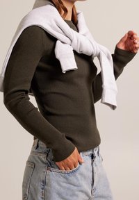 Olivgrön ribbad långärmad tröja med en ljusgrå sweatshirt över axlarna, kombinerad med högmidjade ljusblå jeans.