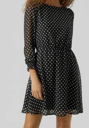 Robe longue noire transparente à manches longues avec des pois blancs, taille élastique et jupe évasée portée par une personne aux cheveux bouclés.
