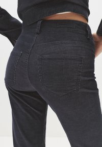 Pogled od blizu na osebo, oblečeno v temnoleve panele hlače z zadnjimi žepi, prikazuje pas in del usklajene jakne.