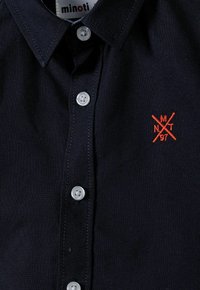 Camicia di cotone blu navy con colletto classico, bottoni bianchi e logo rosso ricamato con linee incrociate e numeri.