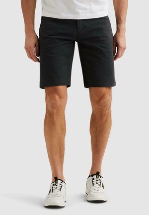 Mężczyzna w czarnych spodenkach do kolan, białych sneakersach z czarnymi i żółtymi akcentami oraz białej koszuli, stojący na białym tle.