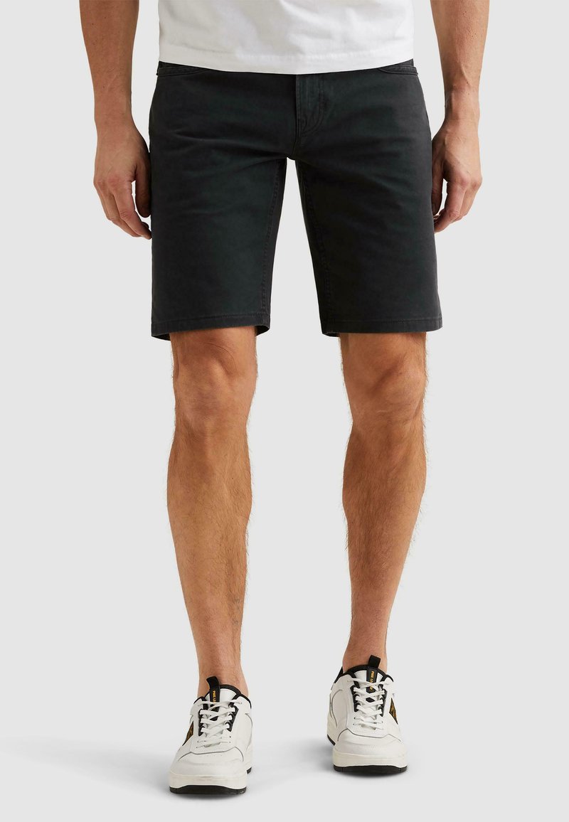 Homme portant un short noir jusqu'aux genoux, des baskets blanches avec des accents noirs et jaunes, et une chemise blanche, debout sur un fond blanc.