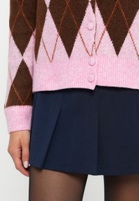 Rudas ir rožinis argyle rašto megztinis su dygsniuotais kraštais ir trimis mygtukais, derinamas su tamsiai mėlyna klostuota sijonu ir permatomomis pėdkelnėmis.