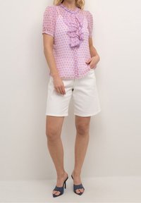 Blusa de color púrpura claro con un patrón de diamantes, frente con volantes y mangas cortas abullonadas. Combinada con pantalones cortos blancos y mule de tacón alto azules.