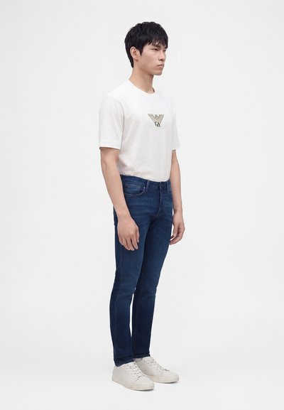 Νεαρός άνδρας στέκεται φορώντας λευκό t-shirt με λογότυπο, σκουρό μπλε τζίν και λευκά αθλητικά παπούτσια σε απλό ανοιχτό φόντο.