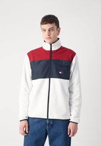 Giacca in pile con una parte superiore rossa, pannello centrale blu navy e parte inferiore bianca. Presenta una chiusura con zip e una tasca laterale con dettaglio a bottone.