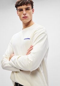 Langärmliges cremefarbenes T-Shirt aus Baumwolle, mit lila Logo auf der Brust und lockerer Passform mit leicht schmaler zulaufenden Ärmeln.