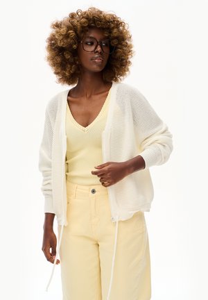 Kobieta z kręconymi włosami nosząca duże okulary, kremowy sweter na guziki, żółty prążkowany top i jasnobeżowe spodnie z wysokim stanem, patrząca na bok.