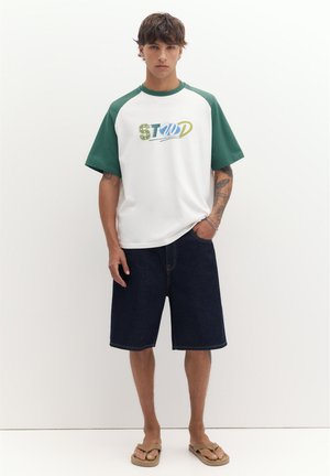 Ung mand iført hvid og grøn kortærmet skjorte med teksten "STWD", mørke denimshorts, tatoveringer og sandaler, stående mod hvid baggrund.