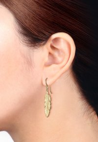 Goldfarbene, blattförmige Ohrringe mit detaillierter Federstruktur und einem Hakenverschluss. Tragen auf dem linken Ohr, gestylt mit glattem braunem Haar.