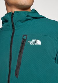 Chaqueta de cremallera en color verde azulado con capucha, que presenta un bolsillo con cremallera negro y un logo blanco en el pecho. Tejido suave y patrón texturizado.
