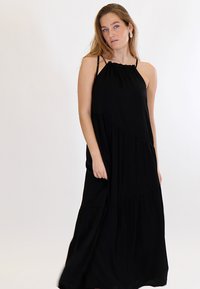 Pikkade juustega naine, kes kannab õhukeste rihmadega musta ilma varrukateta maksikleit tavalise valge tausta ees.