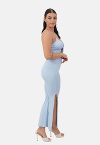 Vestido de color azul claro con detalles de nudo en la parte delantera, aberturas laterales y una silueta ajustada. Presenta tirantes transparentes y zapatos de tacón stiletto.