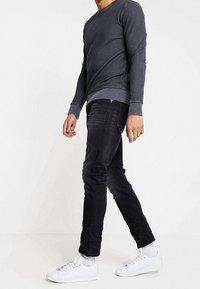 Sudadera de manga larga gris oscuro con cuello redondo, diseño ajustado, combinada con jeans negros de corte slim y zapatillas blancas. Tejido texturizado, acentos mínimos.